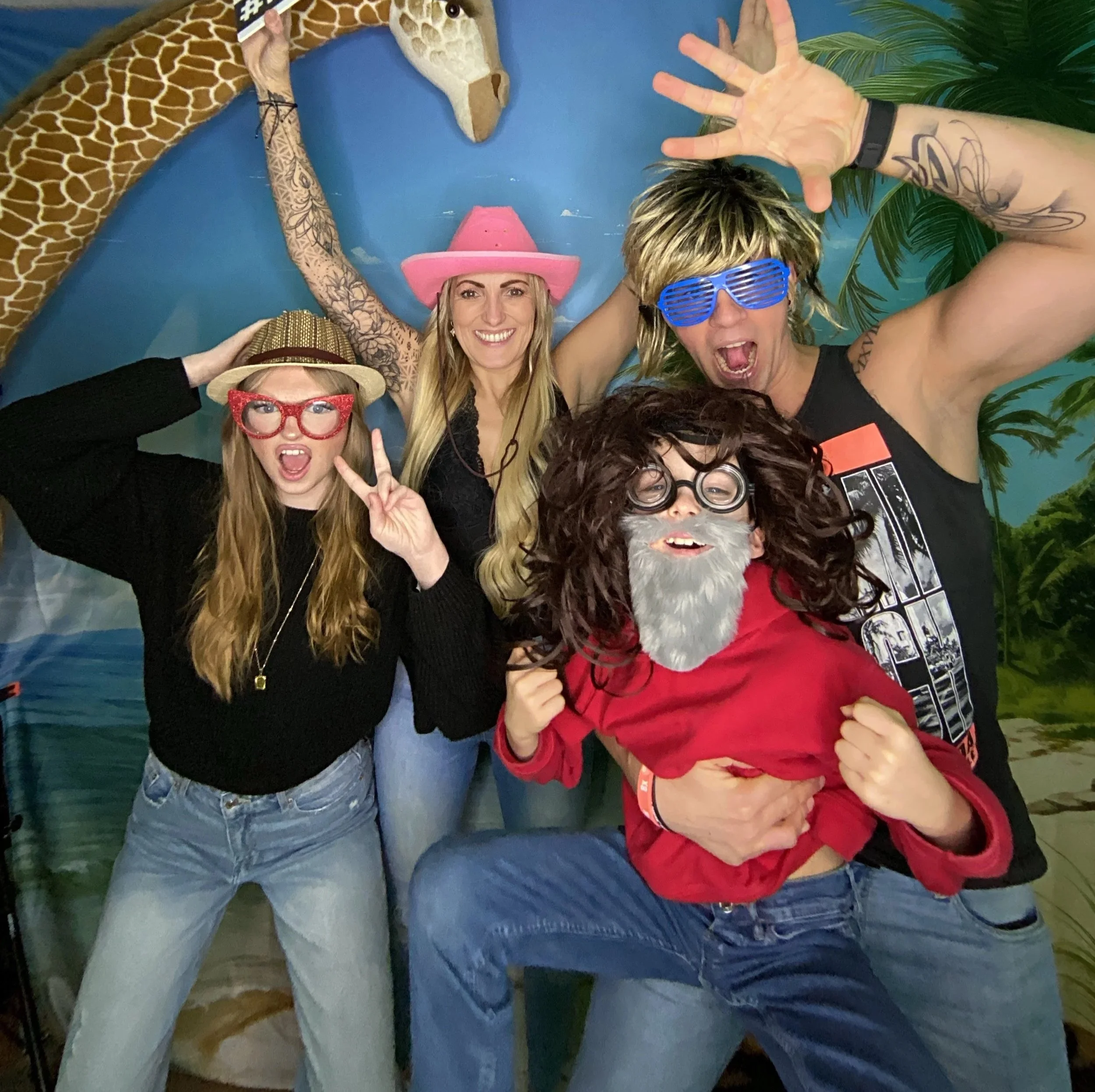 Eine Gruppe von Menschen in einem Raum, die fröhlich in die Kamera lächeln, einige tragen lustige Accessoires wie eine rosa Cowboyhut, eine rote Brille, eine Sonnenbrille und einen roten Bart. Fotobox.