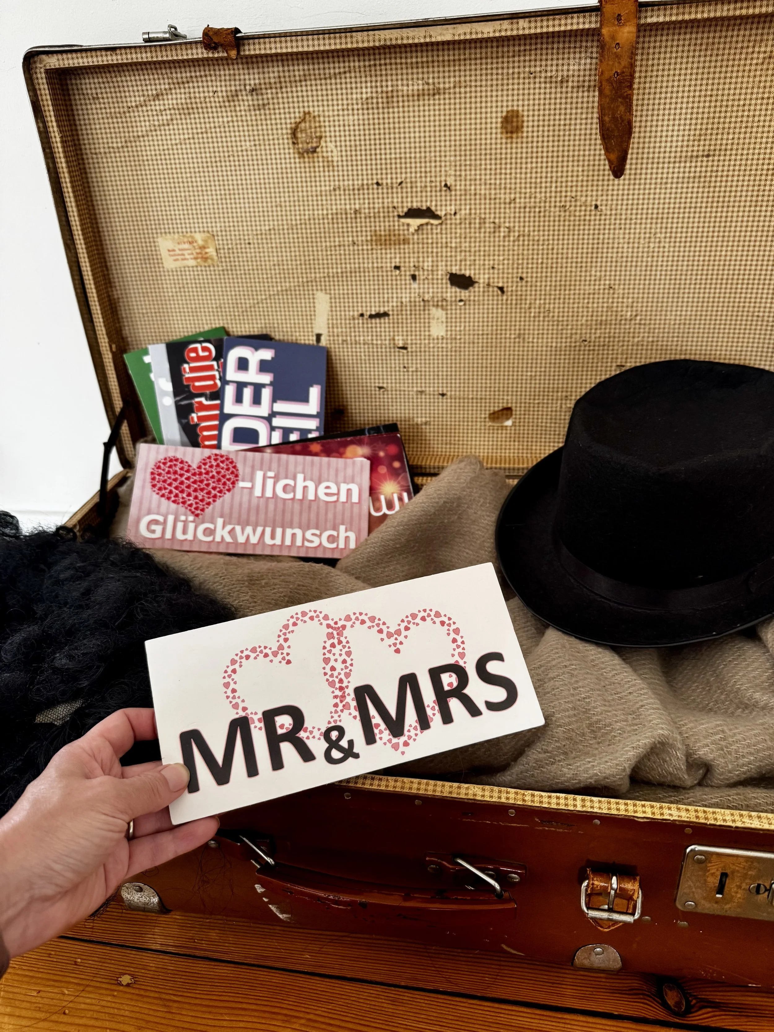 Eine Hand hält ein Schild mit der Aufschrift »MR & MRS«, umgeben von einem Herz aus roten Pünktchen, vor einem Koffer. Im Hintergrund befinden sich verschiedene Hochzeitsdekoartikel und Grußkarten.