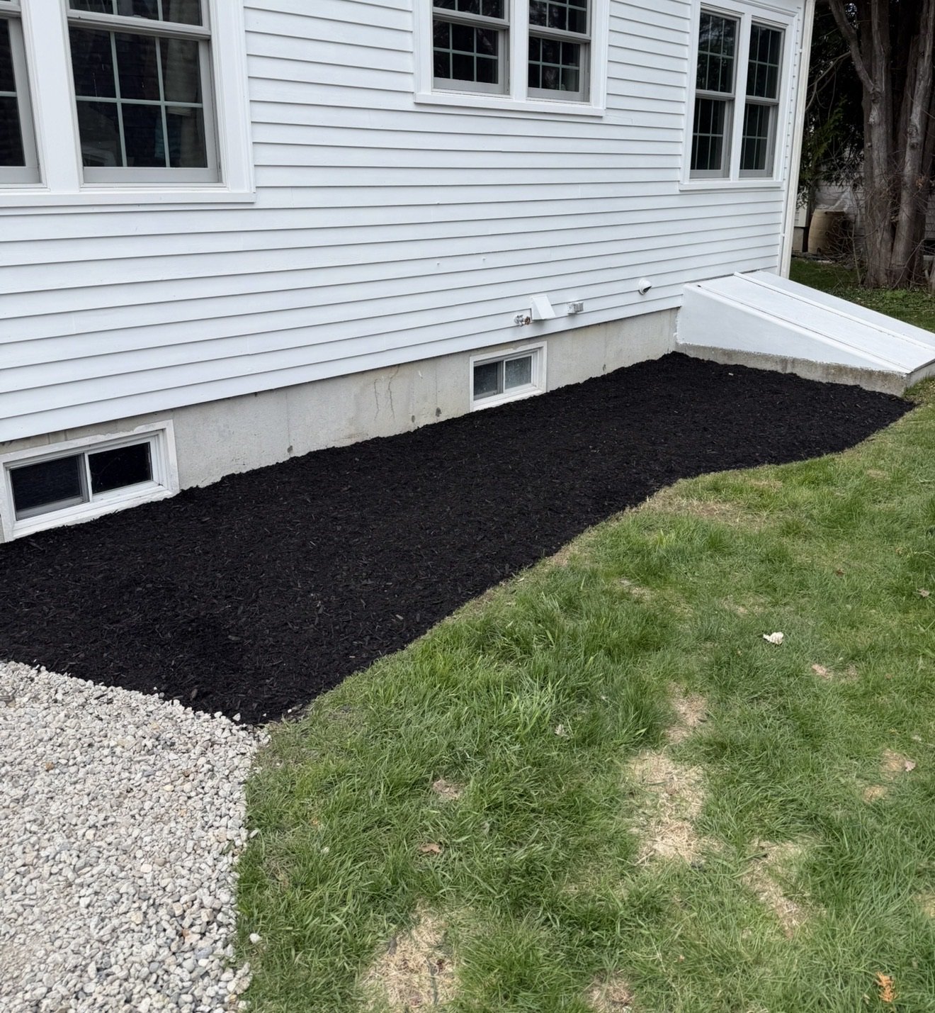 A freshly mulched garden bed next to a house with white siding, small window wells, and a small basement window; the garden bed is edged with white gravel and bordered by a grassy lawn.