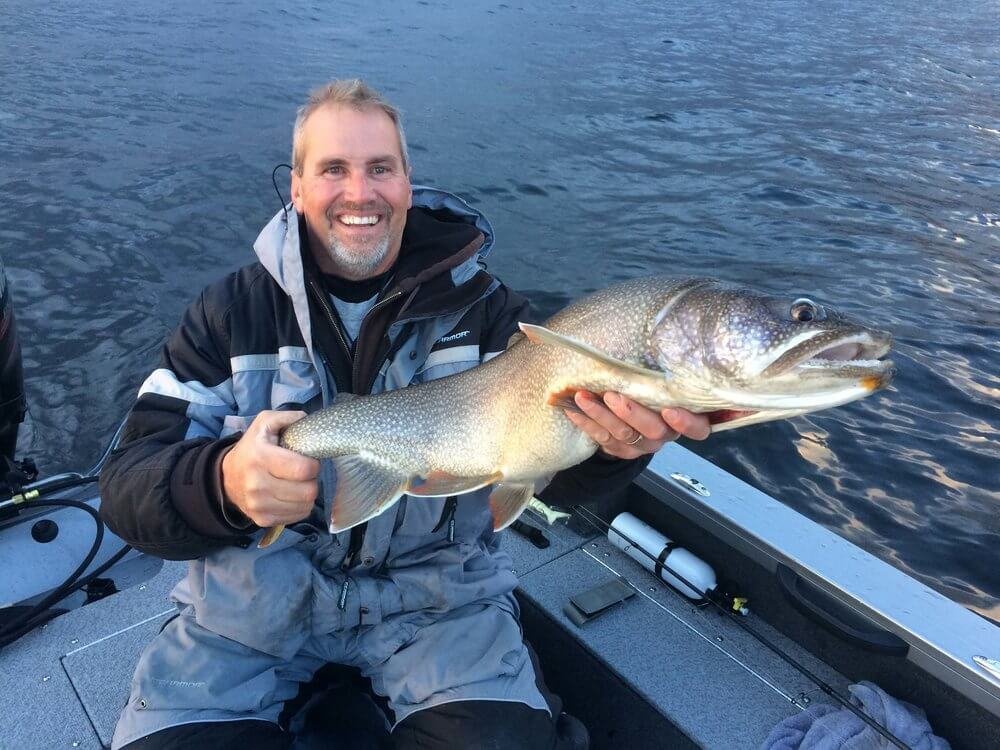 Man in a gray and black fishing jacket sitting on a boat, smiling, holding a large fish, likely a pike or muskie, over water.