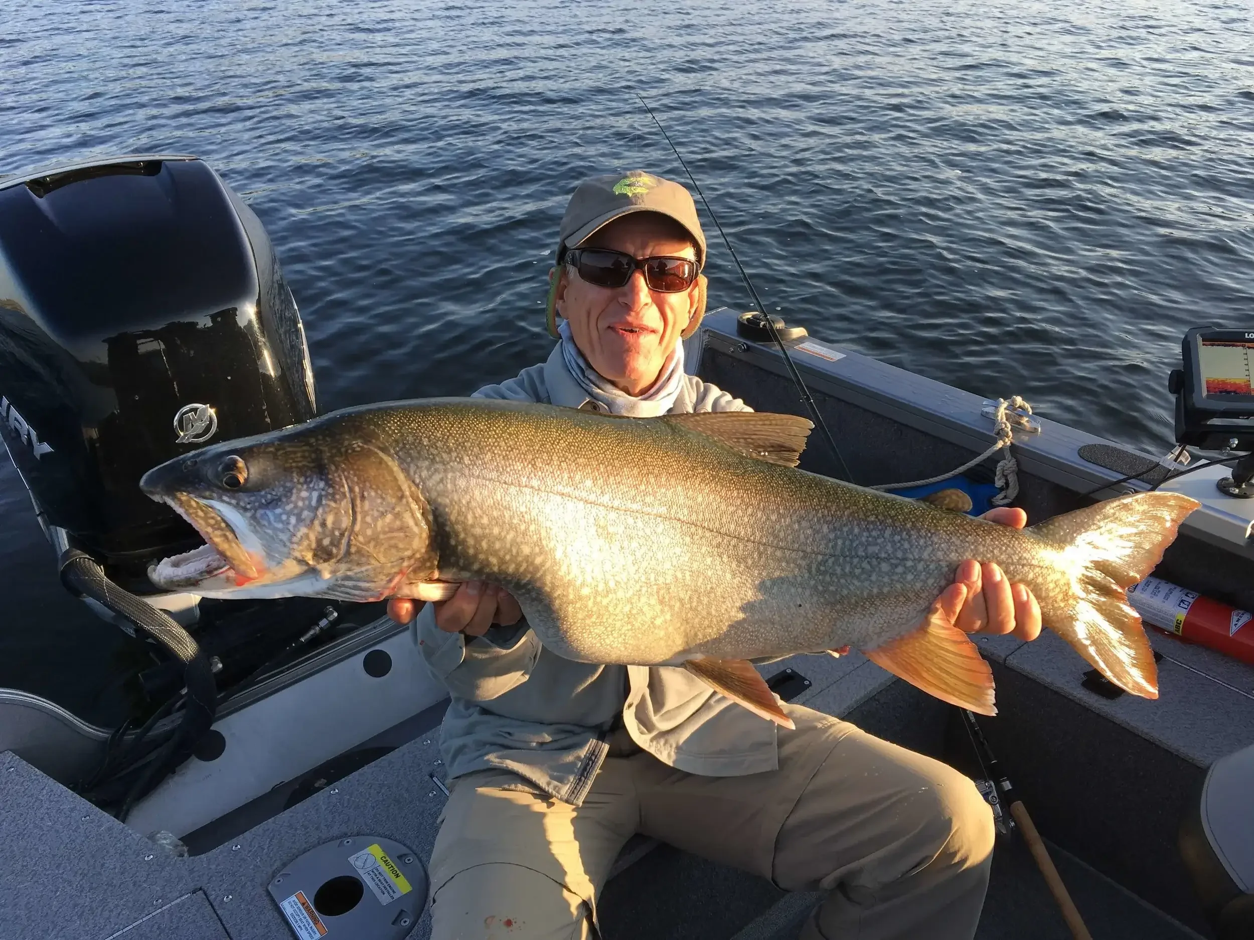 A man on a boat holding a large fish he caught while fishing on open water during sunset.