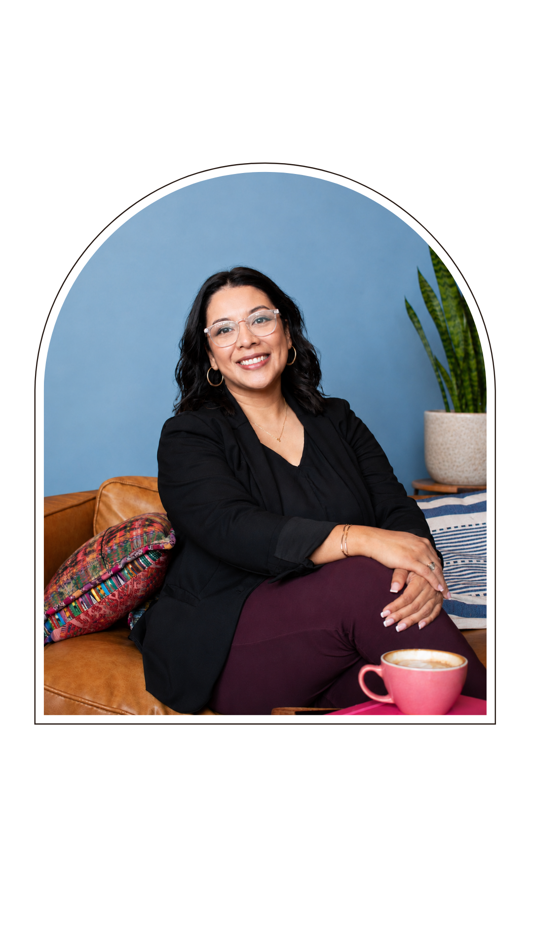 Smiling woman with glasses and dark wavy hair sitting on a sofa with colorful pillows, wearing a black blazer and maroon pants, a pink mug on a table in front of her, and a potted plant in the background against a blue wall.