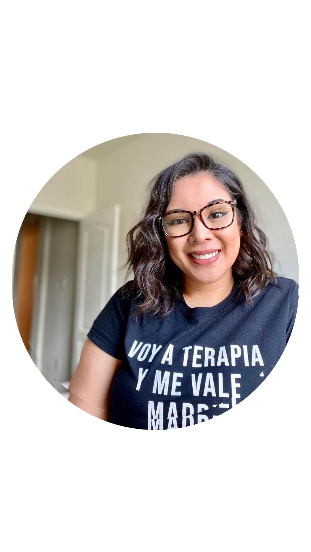 A woman with glasses and curly hair smiling at the camera, wearing a black T-shirt with Spanish text that says, 'Voy a terapia y me vale madre'.