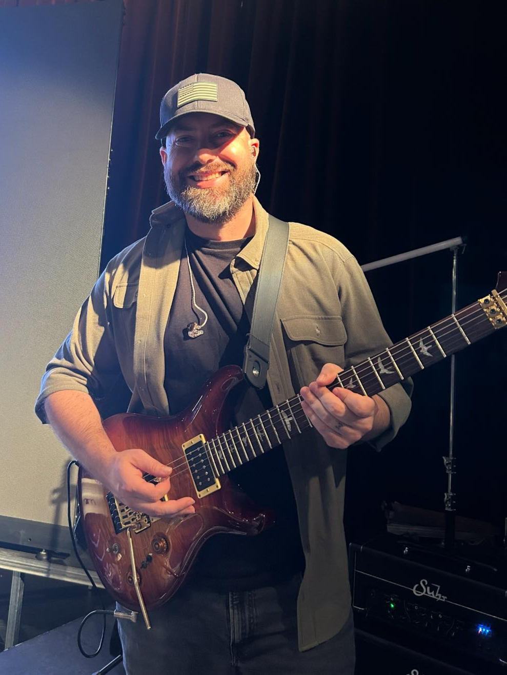 A man with a beard smiling while holding an electric guitar, wearing a cap and casual clothing, in a stage setting with dark background and musical equipment.