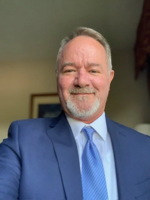 A middle-aged man with gray hair and beard smiling, dressed in a blue suit and tie, taking a selfie indoors.