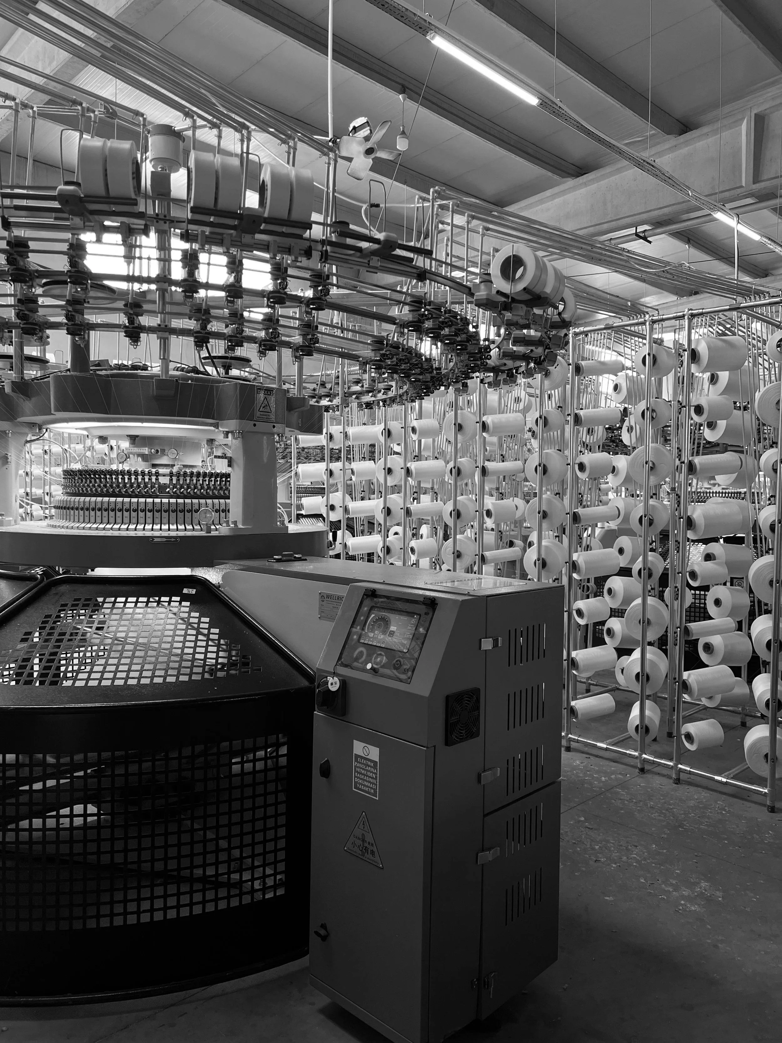 Black and white photo of industrial textile machinery with numerous spools of thread and a control panel in an industrial setting.