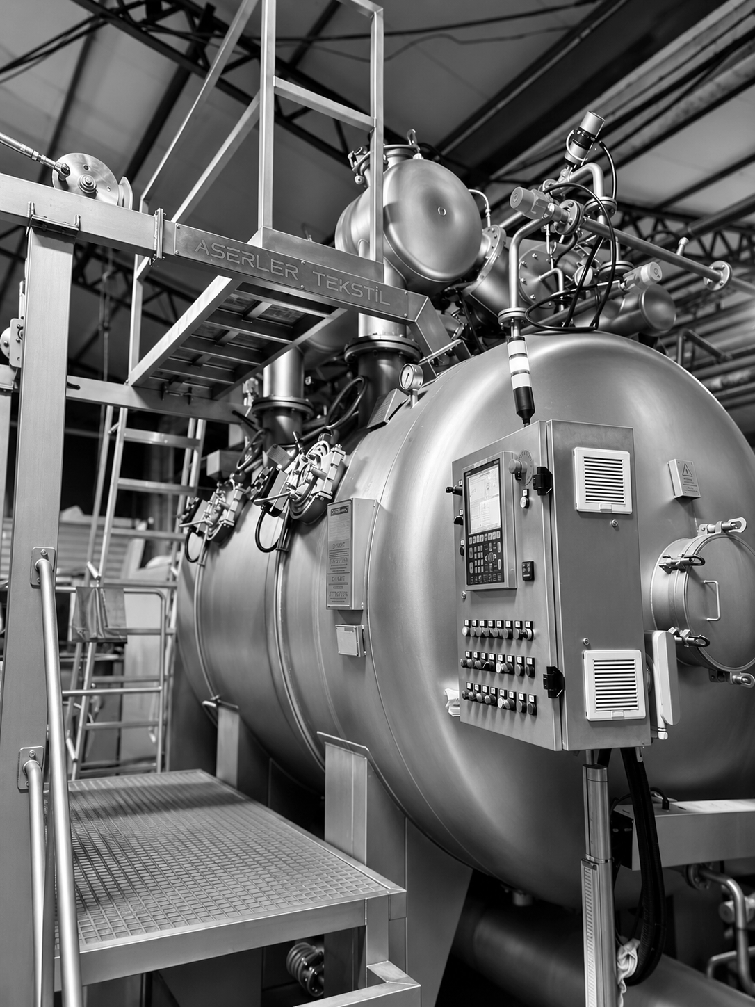 Black and white photo of large industrial equipment, including a cylindrical vessel with control panels and multiple pipes and valves, in a factory setting.