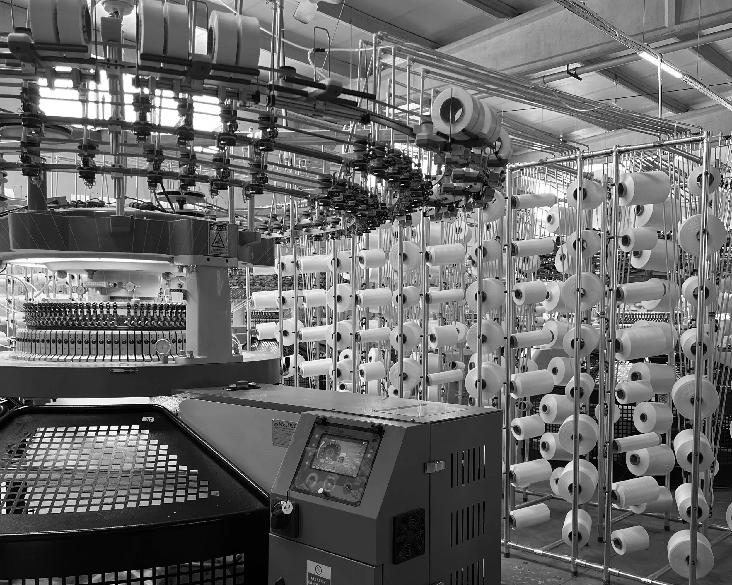 Industrial manufacturing area with numerous white spools of material mounted on metal racks, and control panels with digital displays and electronic components.