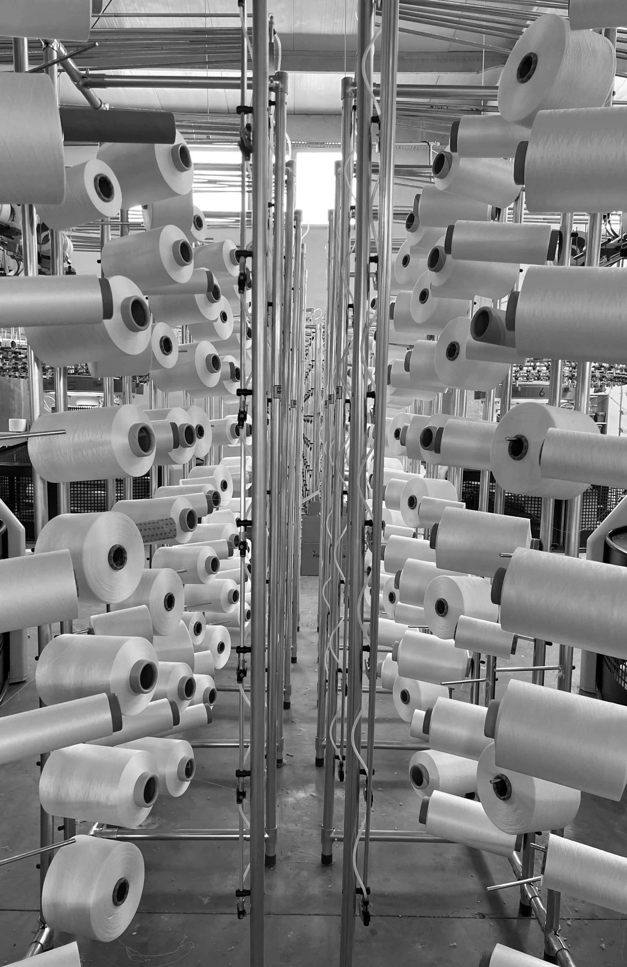 Inside a textile manufacturing facility with multiple rolls of yarn stacked on metal racks, creating a structured and repetitive pattern.