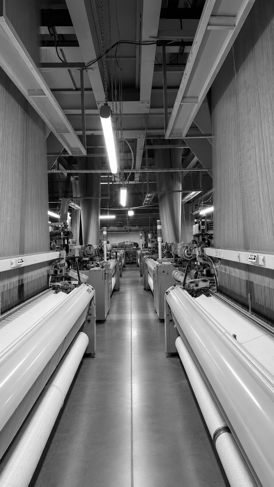 A black and white photograph of a manufacturing or industrial facility with large machines aligned on both sides of a central aisle, ceiling pipes, and hanging lights.
