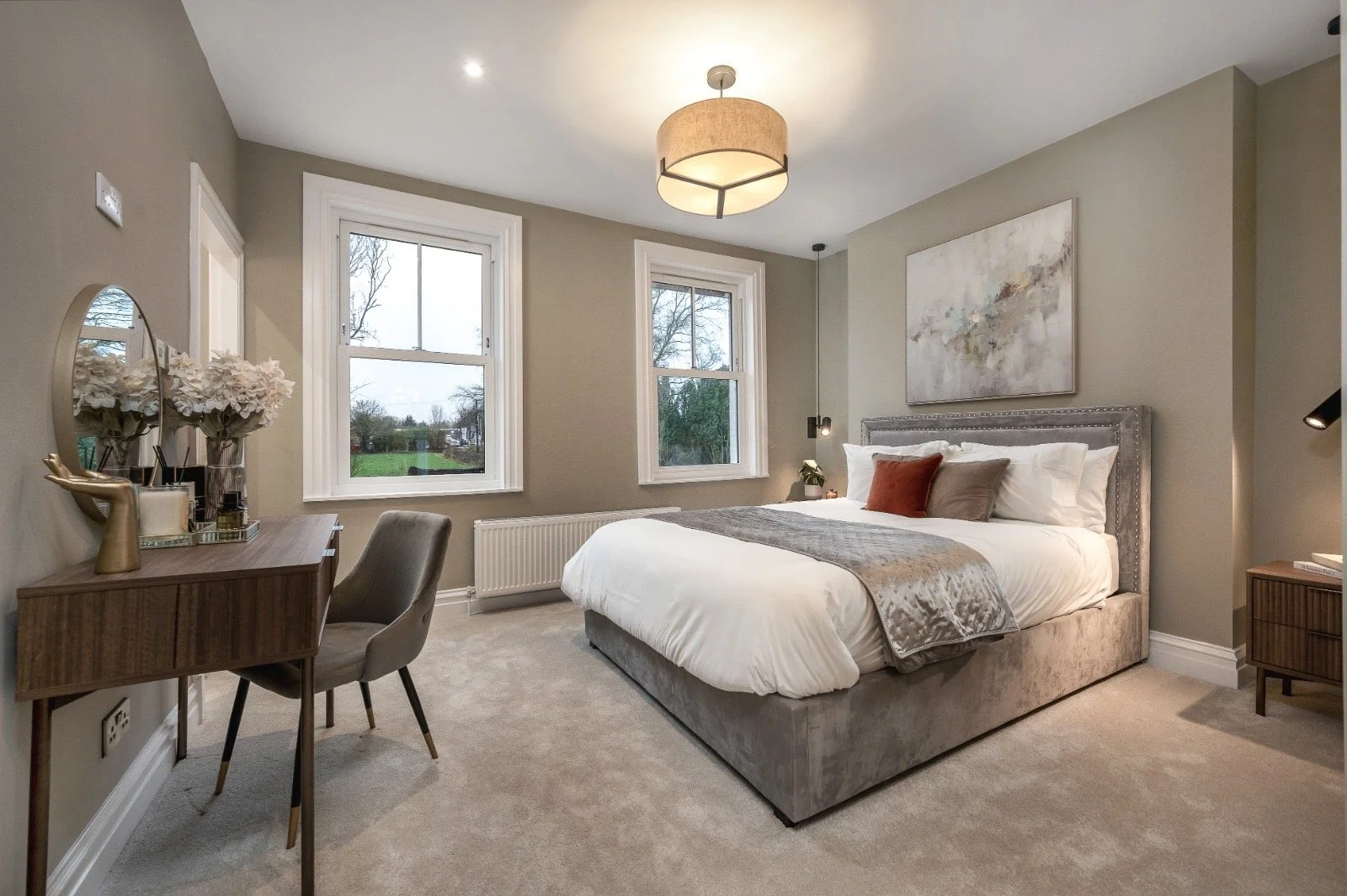 A modern bedroom with a large bed with gray upholstered headboard and white bedding, two windows with views of trees, a wooden desk with flowers, a gray chair, and abstract wall art.