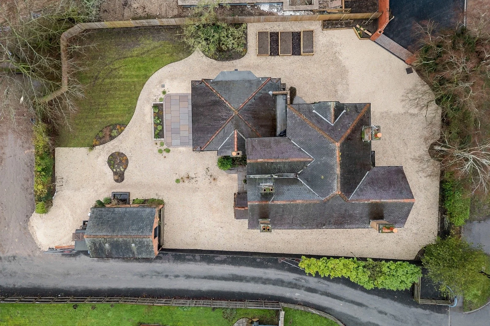 An aerial view of a large house with multiple sections, surrounded by a combination of gravel yard, garden beds, trees, and a driveway. There is a green grassy area at the top and to the left of the house. The house roof is dark with several chimneys