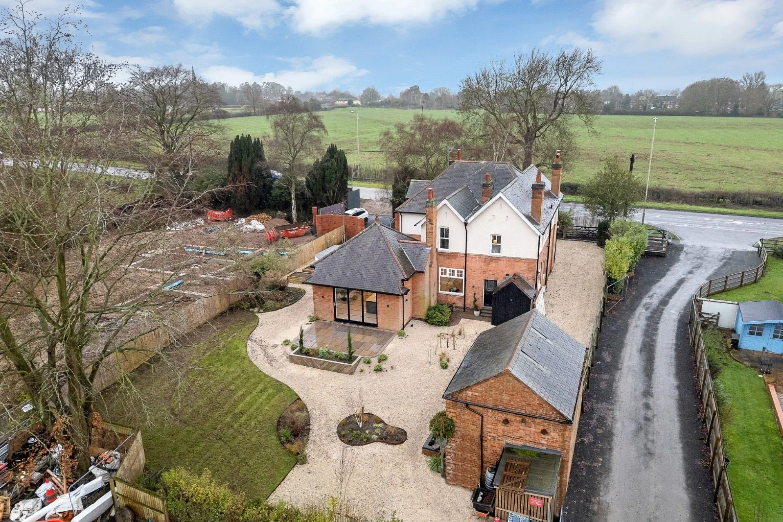 Aerial view of a large white and brick house with multiple chimneys, surrounding a landscaped yard with a gravel path, small garden beds, and near a curved asphalt driveway, set in a green, rural landscape with trees and open fields in the background.