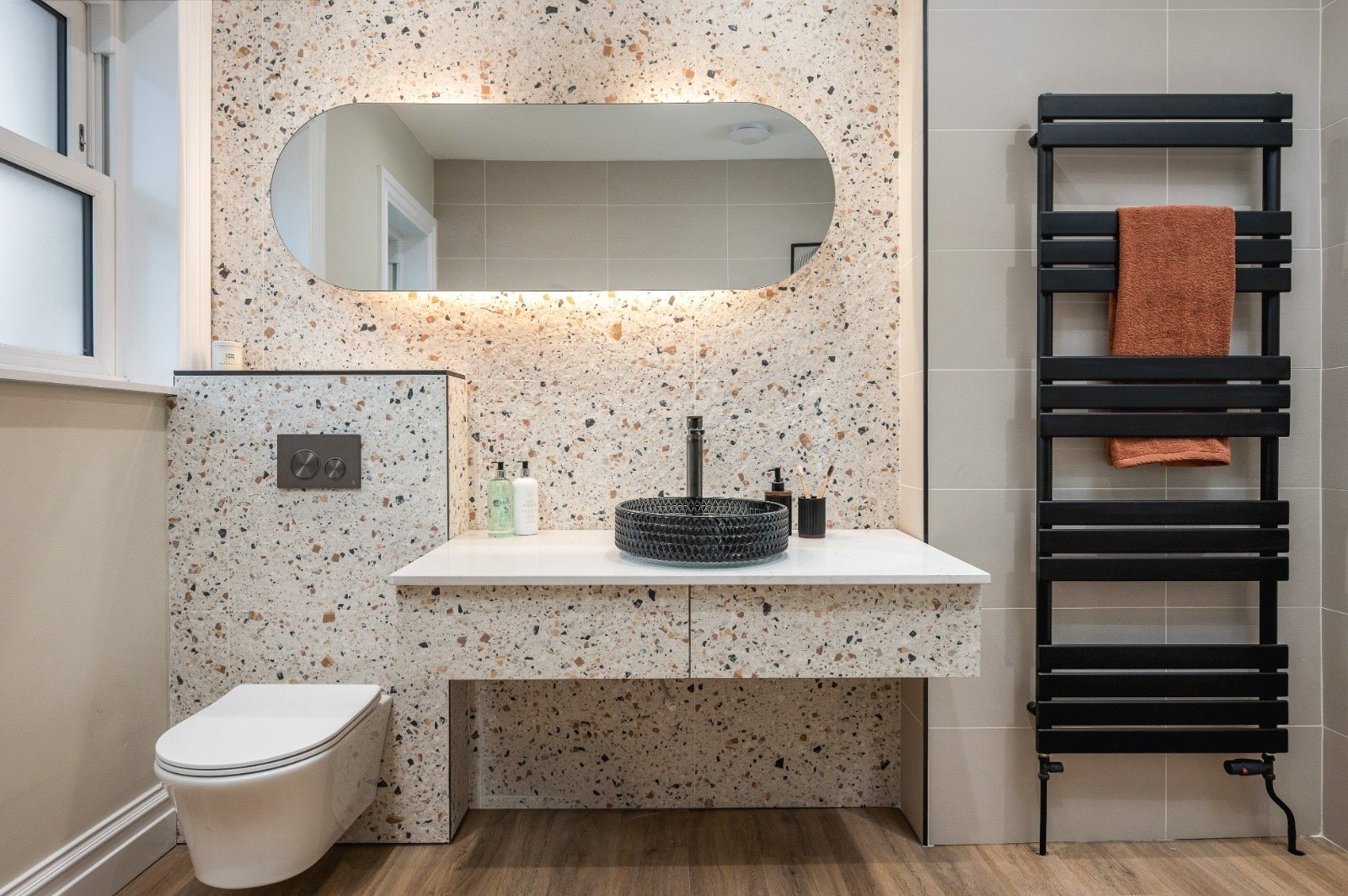 Modern bathroom with a terrazzo wall, a oval mirror, black sink bowl, white vanity, toilet, and black towel radiator with orange towels.