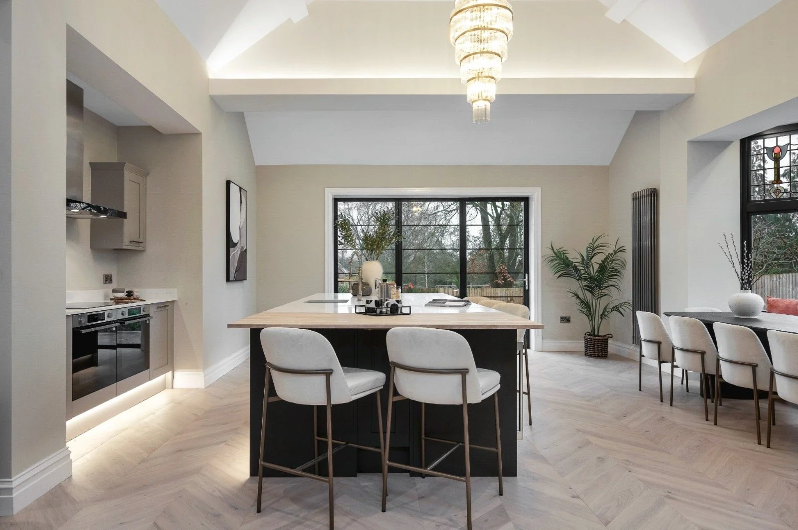 Modern kitchen with a large island, four white chairs, a dining table with six chairs, a large window, and a sliding door overlooking trees.