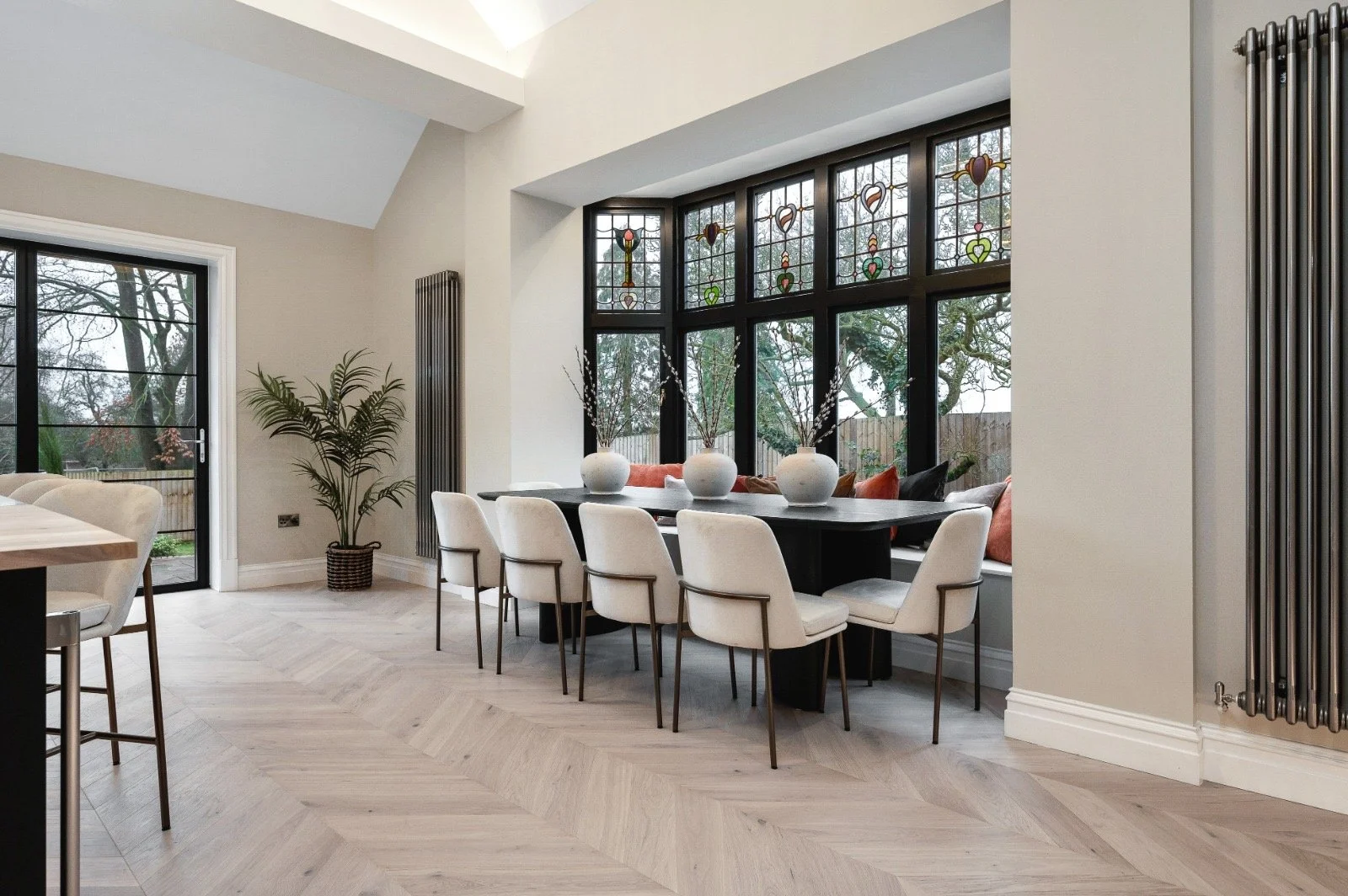 Modern dining room with large black-framed windows and a built-in window seat with cushions, white walls, light wood herringbone floor, and a black dining table with white chairs and decorative vases.