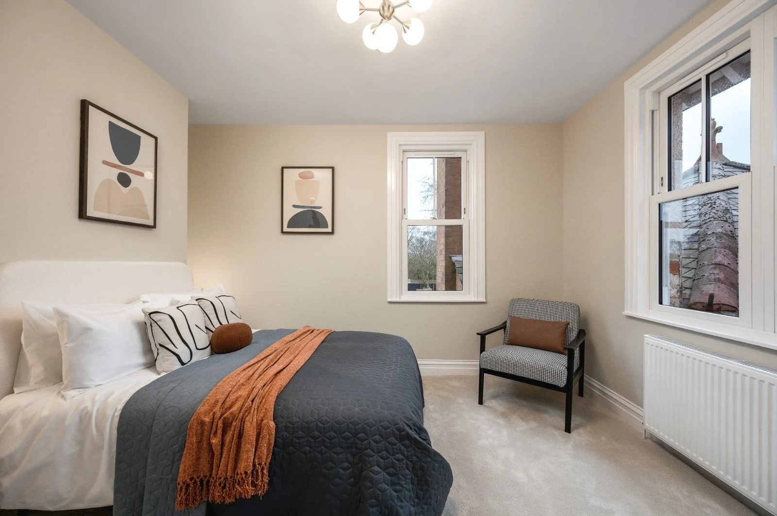A bedroom with a bed featuring white pillows, black and white pillows, and a brown decorative pillow, covered with a dark gray quilt and an orange throw blanket. The room has two abstract art pieces on the beige walls, a window overlooking a brick bu