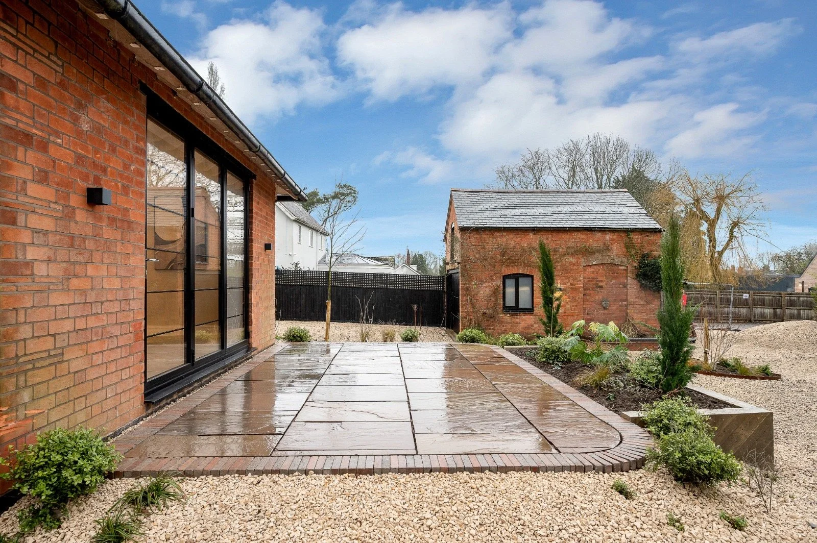 A modern backyard with a wet wooden deck, small plants, and a garden area. A brick house with large glass sliding doors is on the left, and a small brick building with a window is in the background. The yard is enclosed by a black wooden fence, and t