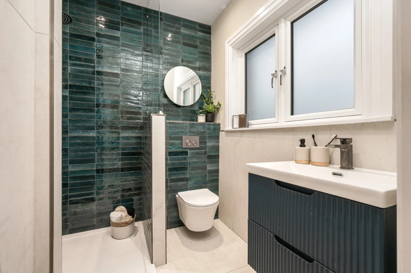 Modern bathroom with a walk-in shower featuring dark green tiles, a wall-mounted toilet, a white sink with a dark blue vanity, and frosted windows.