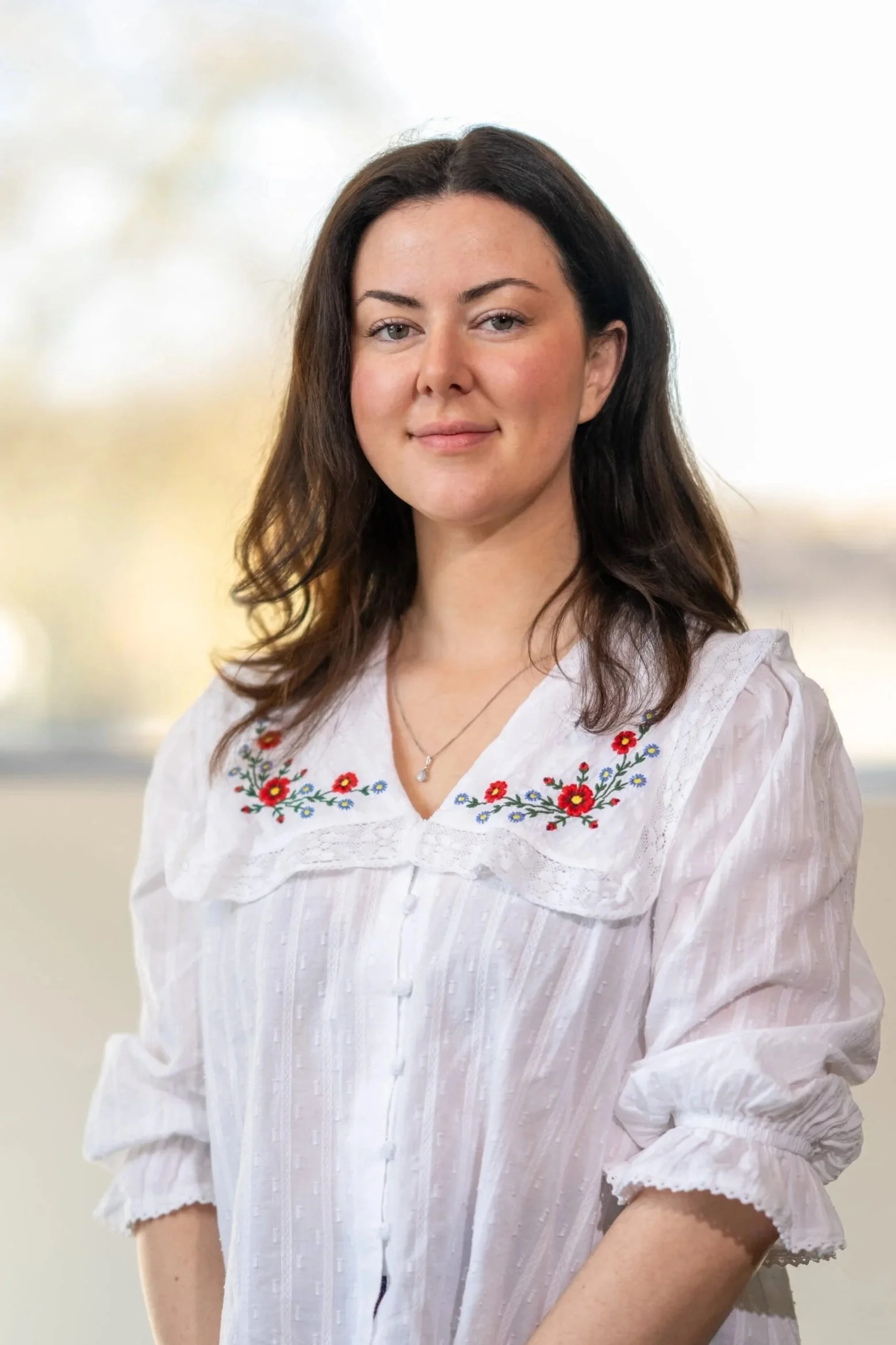 A woman with dark brown hair and light skin wearing a white blouse with colorful floral embroidery around the collar.