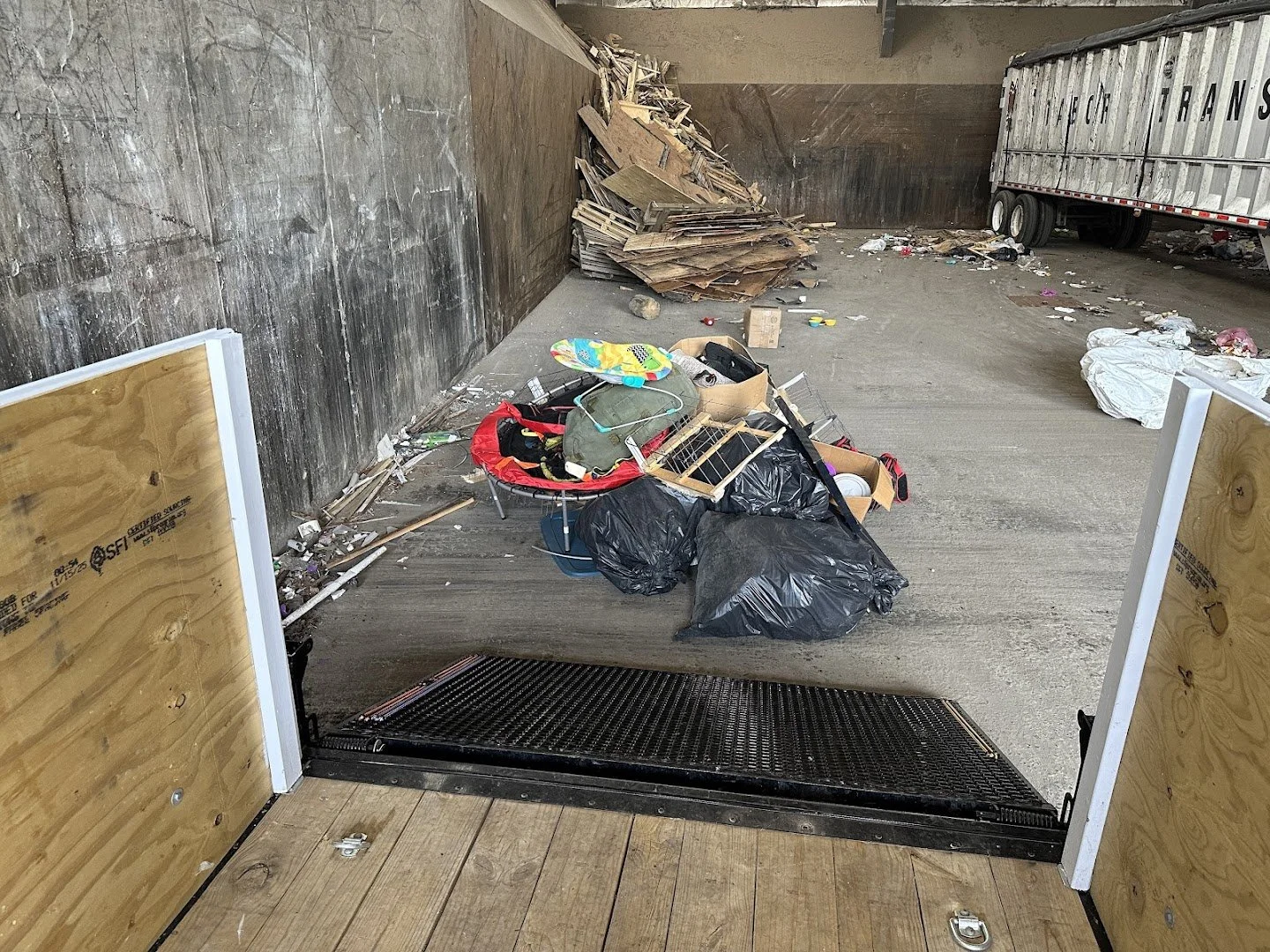 The image depicts the inside of a trailer with its doors open, revealing a pile of trash and debris, including trash bags, broken wood, and discarded items, scattered on the ground just outside the trailer.