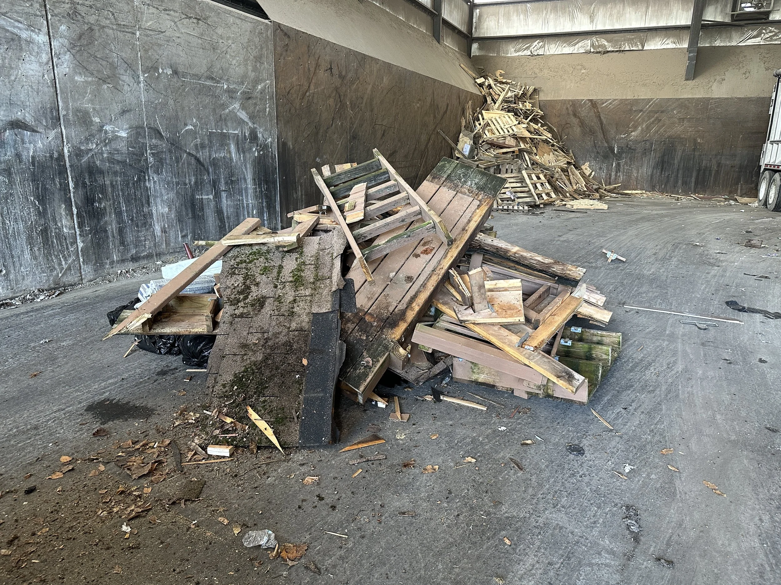 A partially inside view of a construction site with wood pallets and debris piled up on a concrete floor, surrounded by unfinished walls and an exposed ceiling.