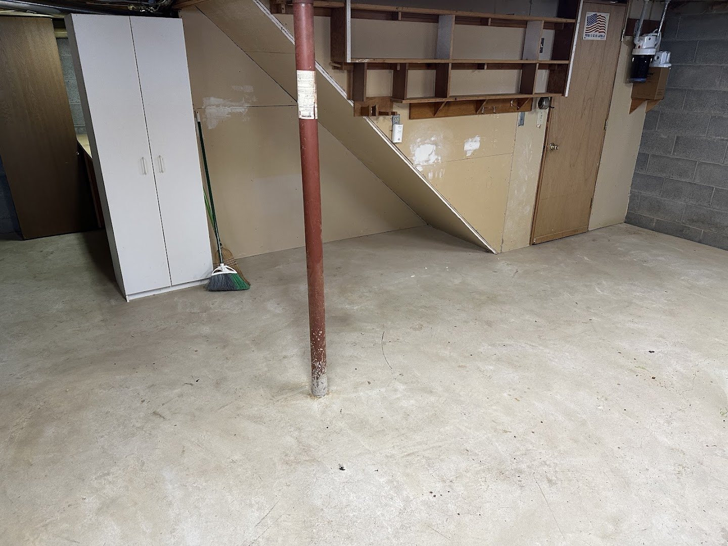 Empty garage with a concrete floor, a white storage cabinet, a broom leaning against the cabinet, a red metal support pole, and a staircase with shelves above. The walls are unfinished with some wood paneling and a gray cinder block wall on the right