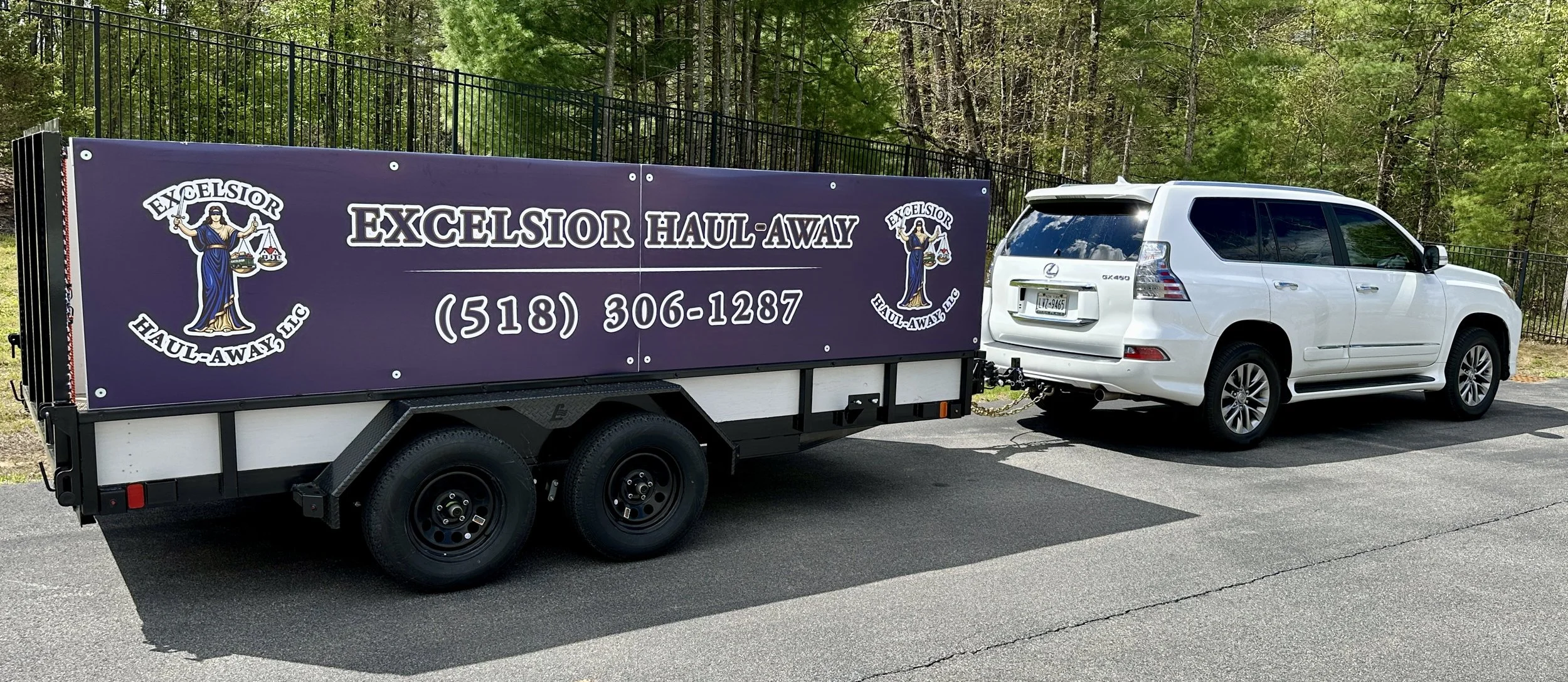 A white Lexus SUV attached to a purple trailer with signage for Excelsior Haul-Away LLC, displaying a logo with a woman holding a sword and scales, and a phone number. The trailer is parked on a paved road with a wooded area in the background.