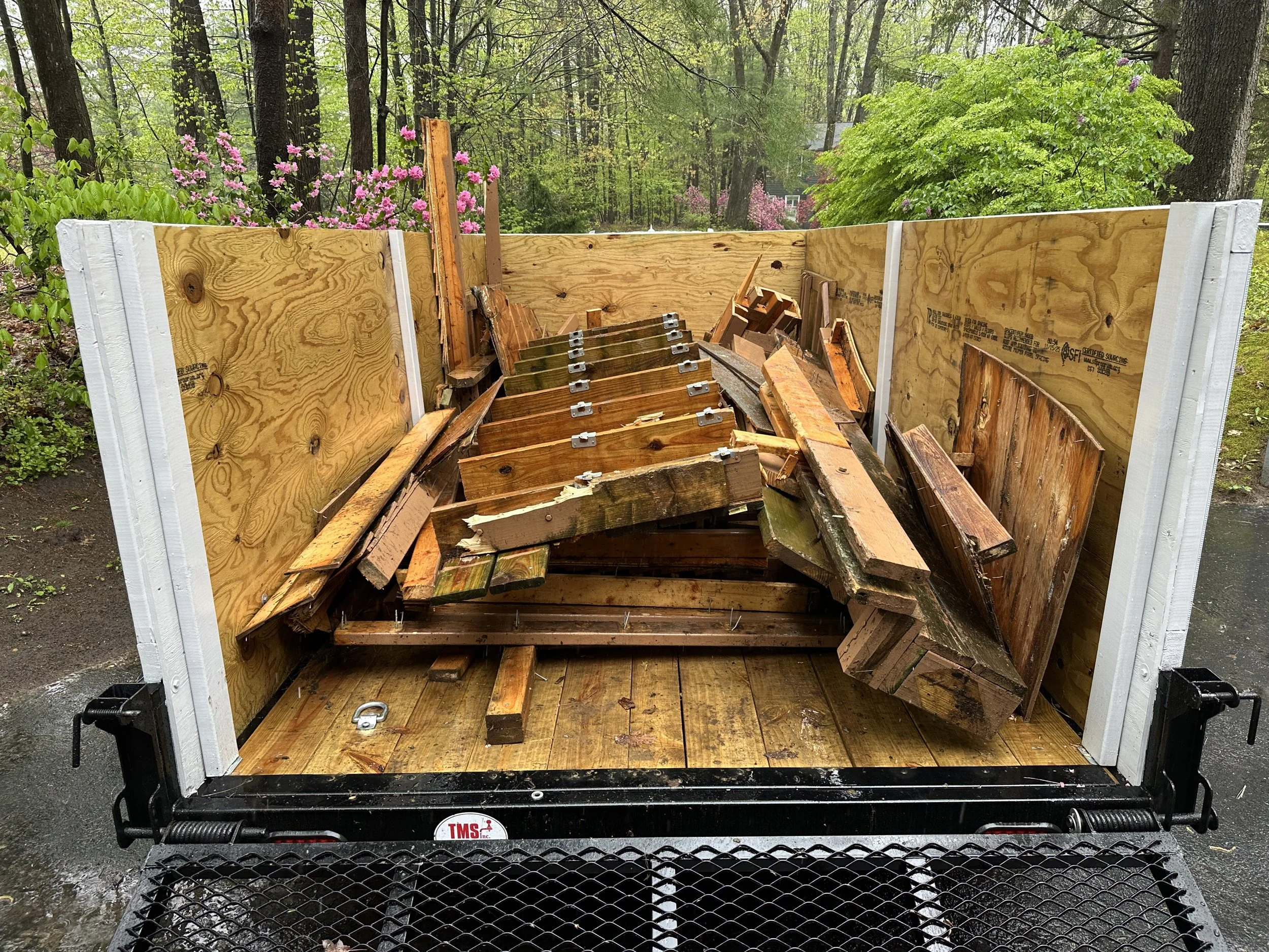 The image shows the inside of an empty truck bed with wooden side panels, which appears to be used for transporting debris or construction materials. It is located outdoors, surrounded by trees and blooming flowers.