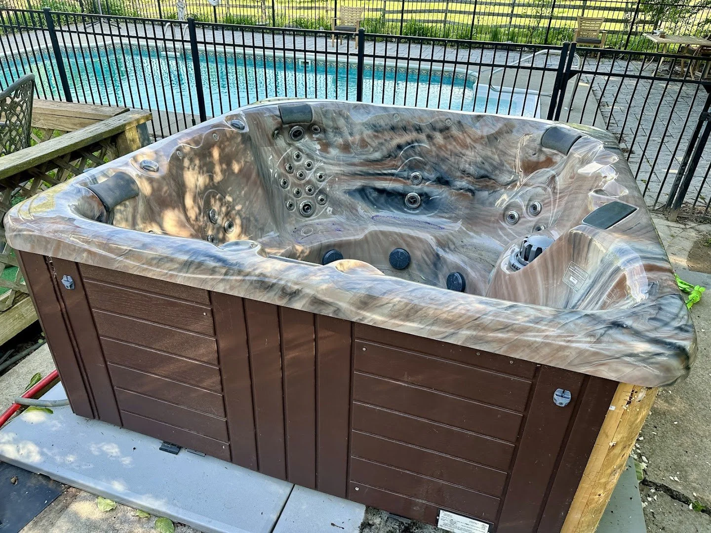 Outdoor hot tub with brown and beige marbled exterior, installed in a fenced backyard next to a swimming pool.