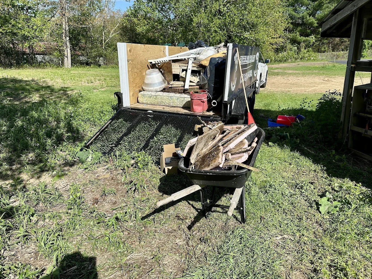 A small work trailer with various tools and materials, including wood planks, a red fuel container, and gardening tools, parked on grass in a backyard with trees and a partially visible wooden structure.