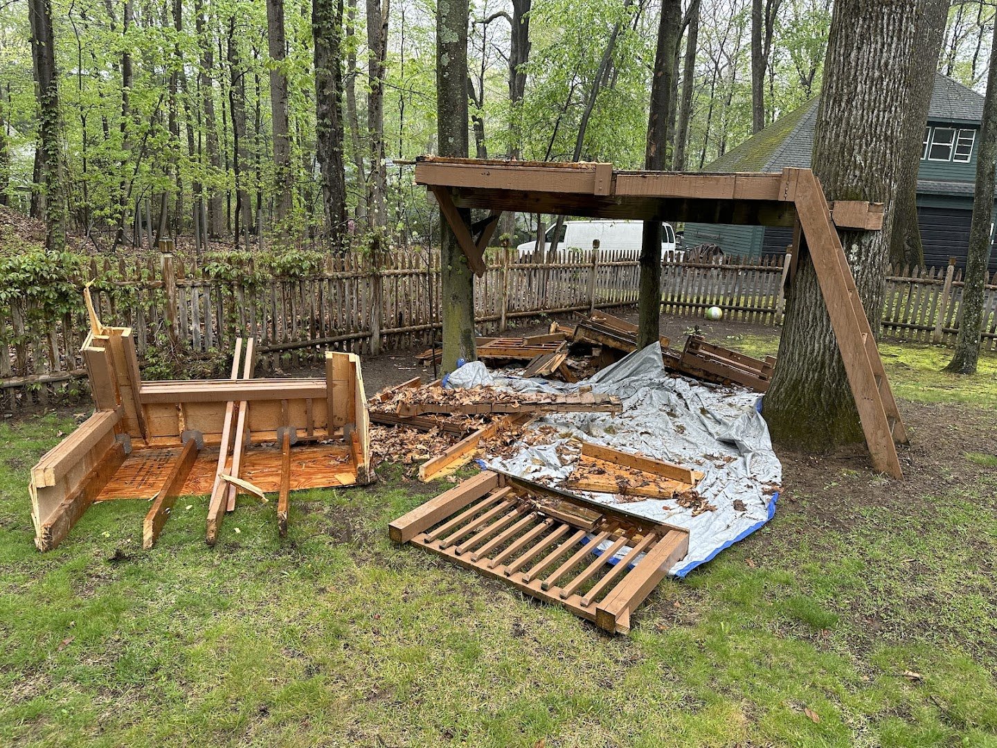 Disassembled outdoor wooden furniture, including parts of a table and slatted pieces, on a grassy backyard with a tarp, trees, and a wooden fence.