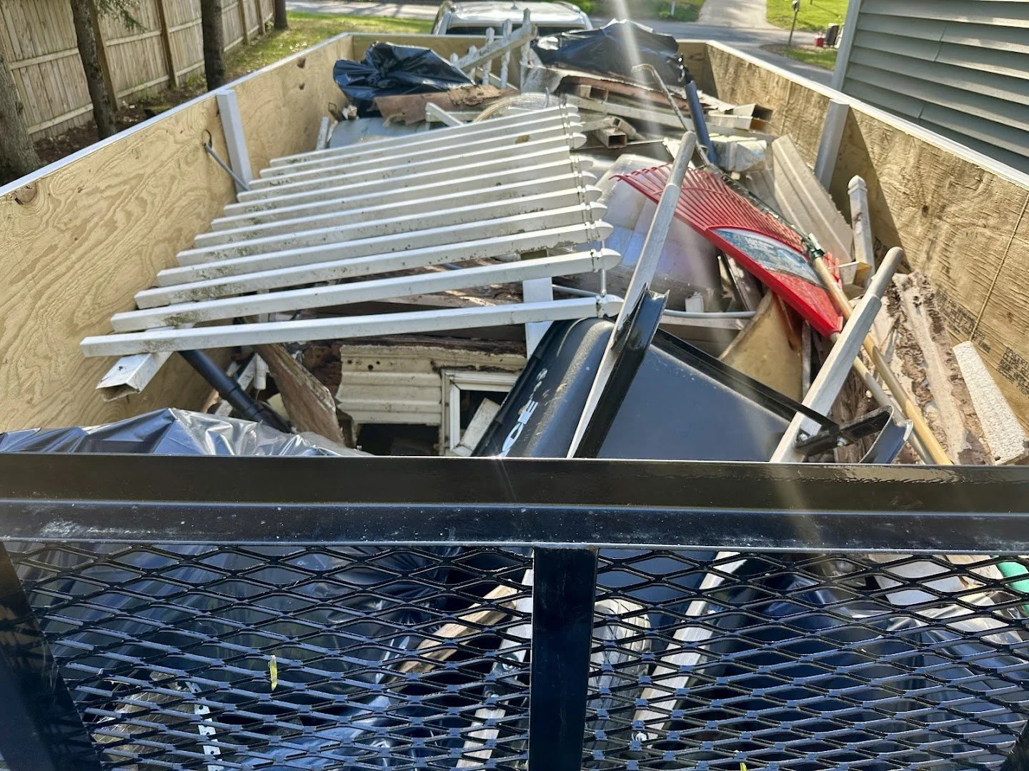 A large black trailer filled with assorted debris including a white metal bed frame, a red car tail light, pieces of wood, metal pipes, and other miscellaneous trash.