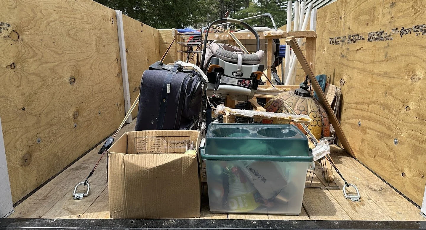 Storage trailer filled with various items including a suitcase, a child's car seat, a large decorative vase, clear plastic storage bin, cardboard box, and various tools and household items.