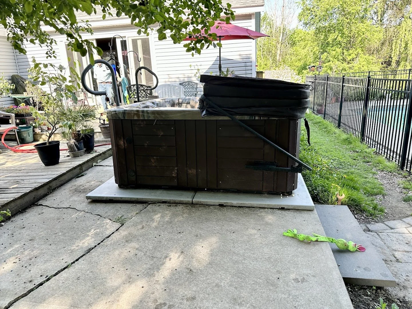 Backyard scene with a hot tub on a concrete pad, surrounded by potted plants, a deck with outdoor furniture, and a black metal fence. An umbrella provides shade, and a child's toy is on the ground nearby. A person stands near a sliding door of the ho