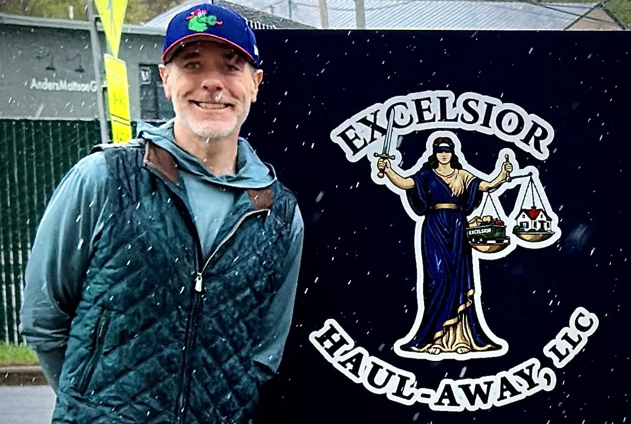 A man with gray hair and a beard, wearing a blue cap with a colorful logo, a gray jacket with a layered vest, and standing in the rain with water droplets visible. He is smiling and standing next to a sign that reads "Excelsior Haul-A-Way Inc," featu