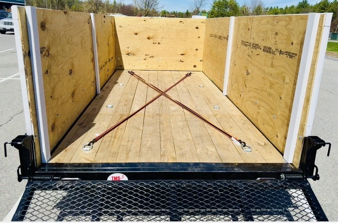 Empty flatbed trailer with wooden side panels and flooring, secured with orange straps, parked outdoors in a lot.