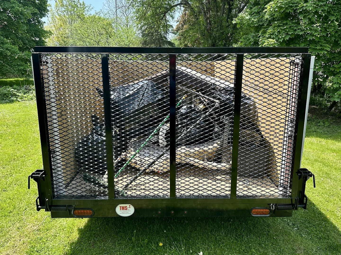 Car engine inside a black metal mesh enclosed trailer on a grassy lawn surrounded by trees.