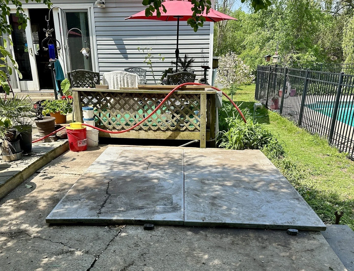 Backyard patio area with a swimming pool, red umbrella, potted plants, and a decked area with chairs, surrounded by a black metal fence, during daytime.