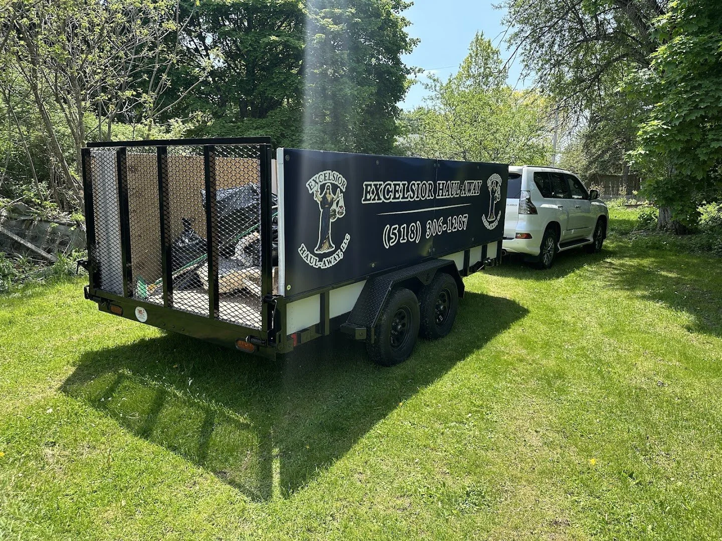White vehicle attached to a black trailer with a sign that reads 'Excelsior Hauling' and a phone number, parked on a grassy area surrounded by trees.