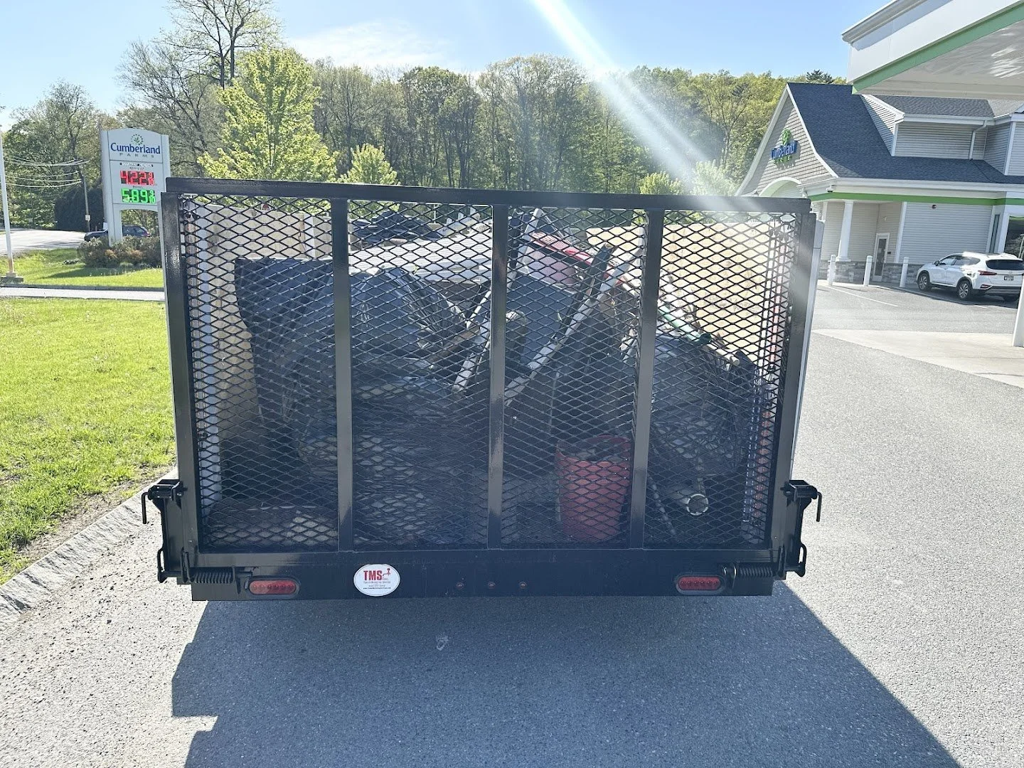 Back of a black utility trailer filled with bicycles, parked in a lot near a green lawn and a Cumberland Farms store with fuel prices displayed.