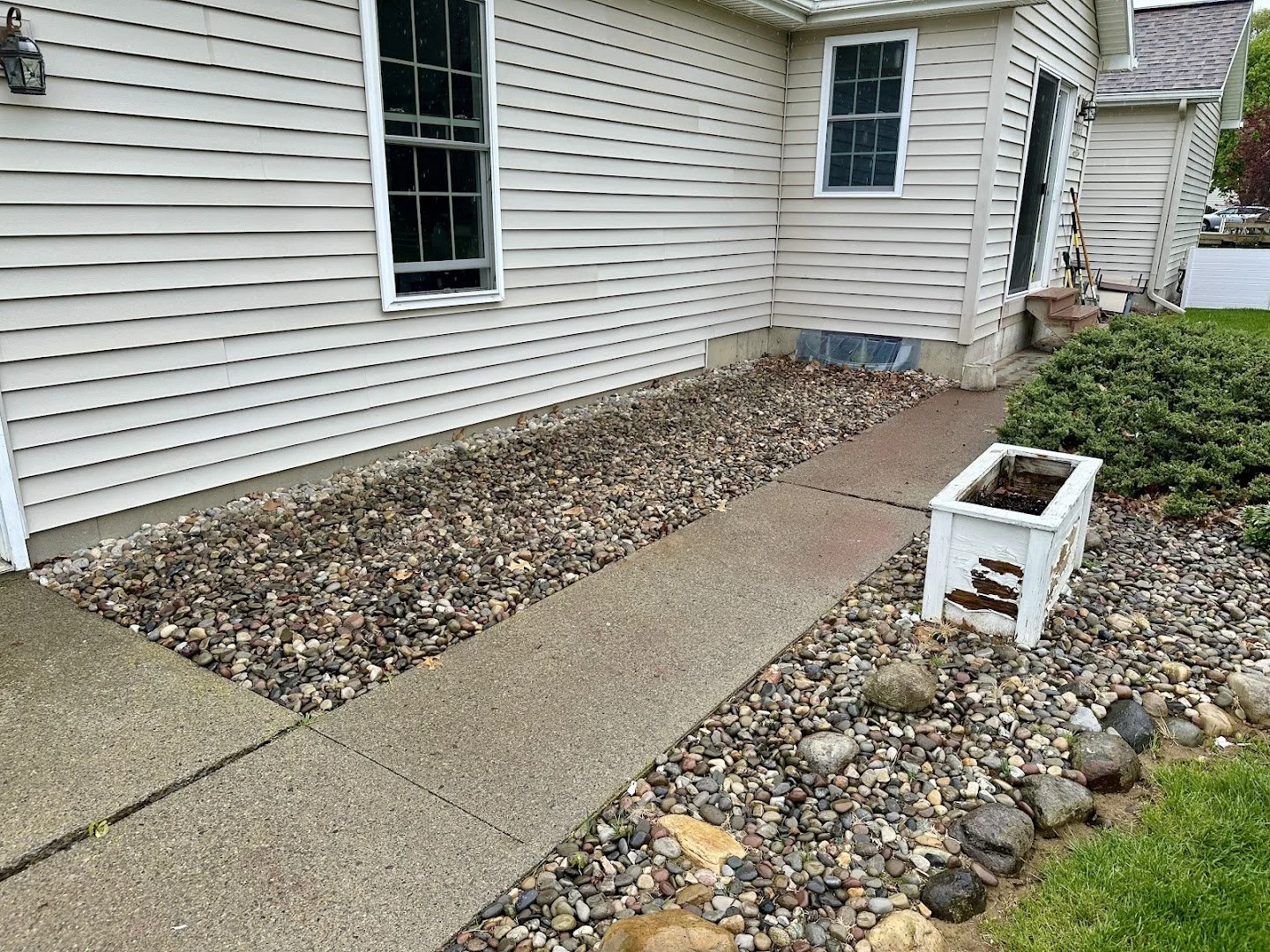 Backyard with concrete pathway, pebble landscaping, a small white wooden planter box, and a house with beige siding and multiple windows.
