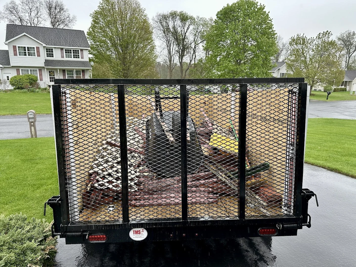 A flatbed trailer parked on a street in a residential neighborhood with a wooden deck and leaves inside, surrounded by green lawns and houses with trees.