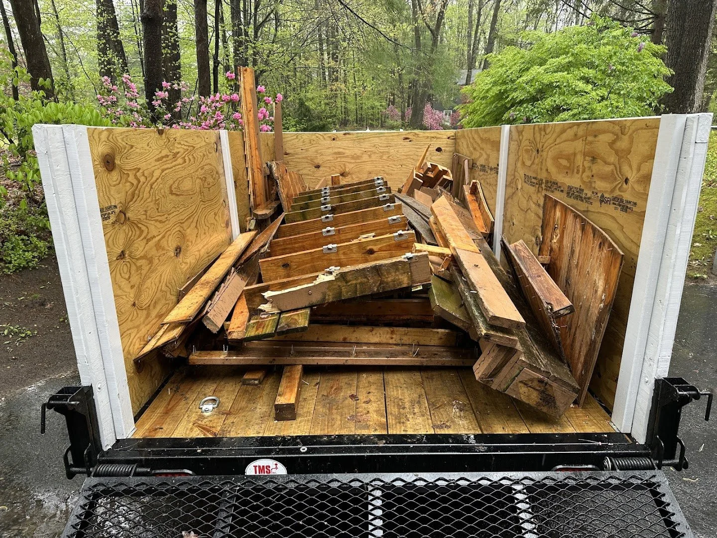 The image shows a flatbed trailer loaded with broken and damaged wooden furniture and debris, surrounded by trees and greenery in an outdoor setting.