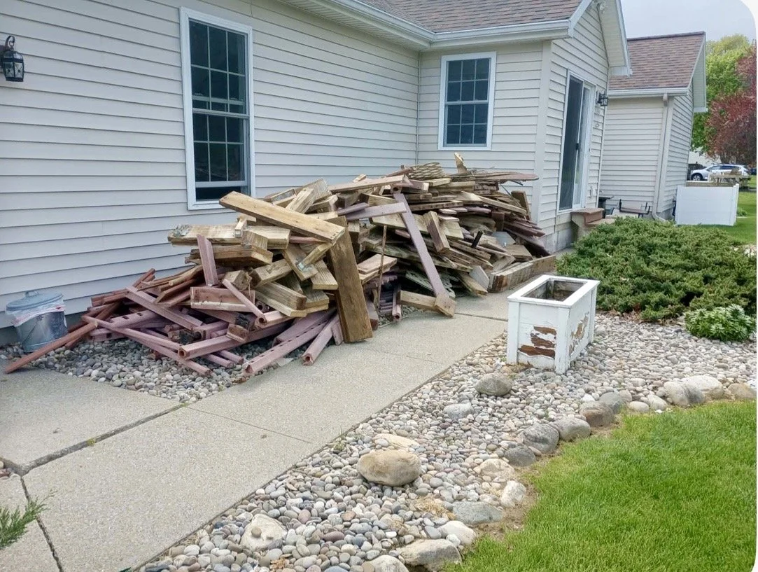 Pile of wooden planks and debris on a concrete sidewalk next to a beige house with stone landscaping and a white planter box.