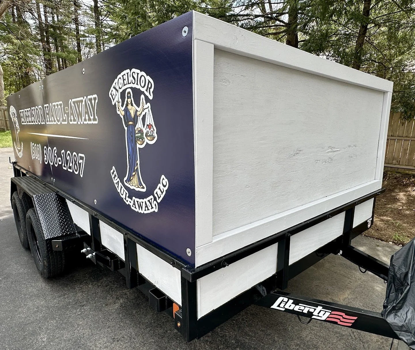 A trailer with a sign that reads "Excelsior Haul Away" and a logo featuring Lady Justice holding scales. The trailer is parked on a driveway with a wooden fence and trees in the background.