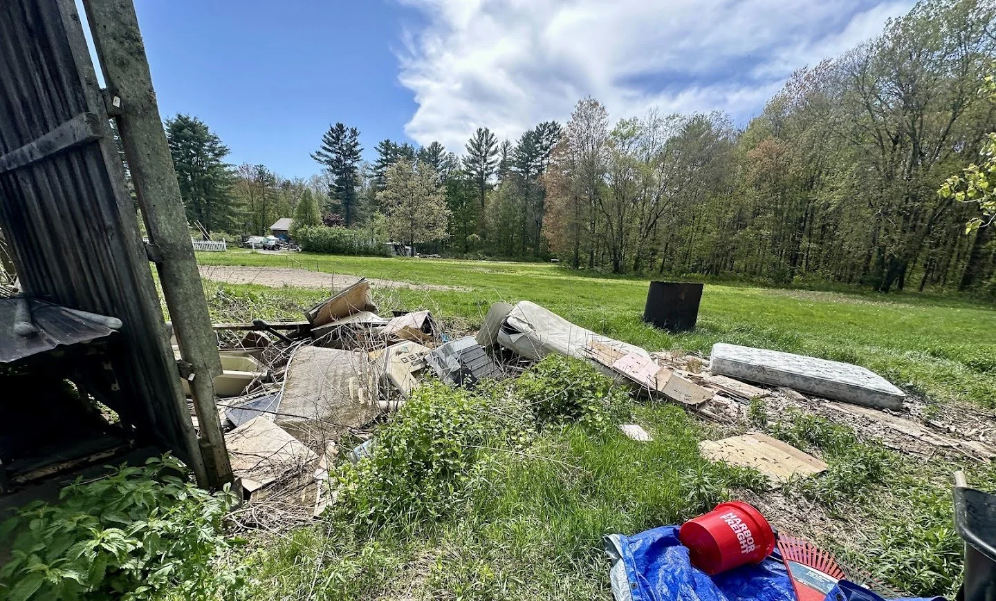 Litter and debris scattered across a grassy area near trees, including trash cans, cardboard, and plastic items under a partly cloudy sky.