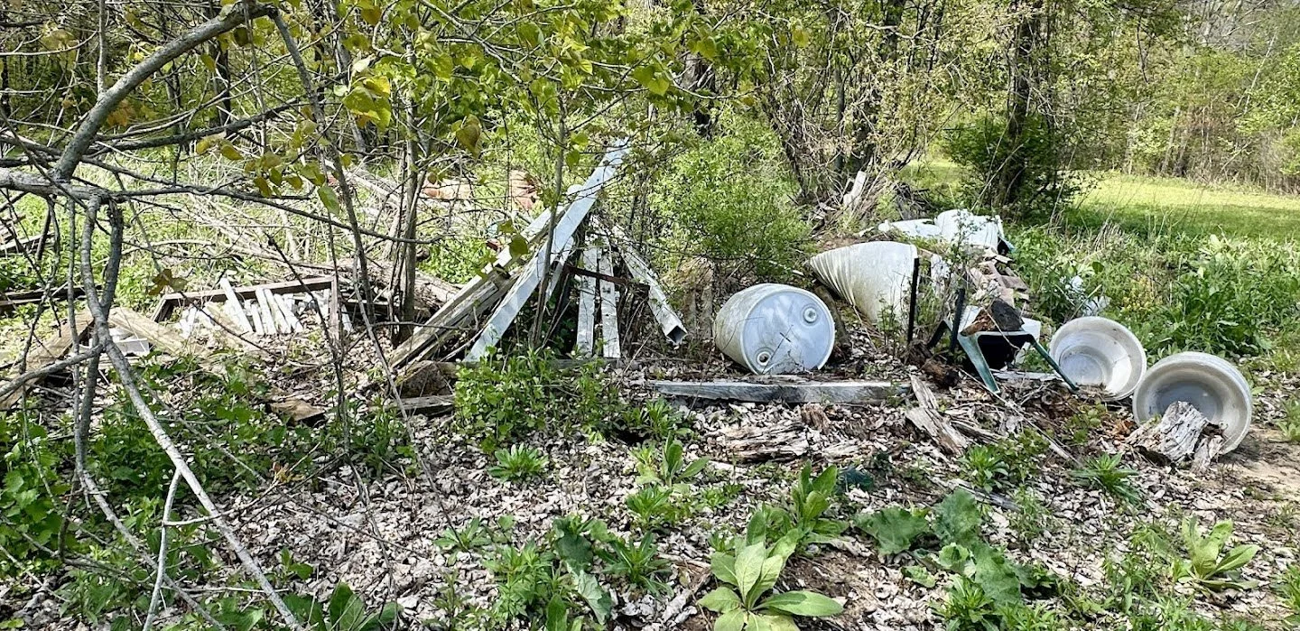 Litter and debris, including metal drums, plastic buckets, and wooden planks, scattered on the ground among bushes and trees in a natural outdoor setting.