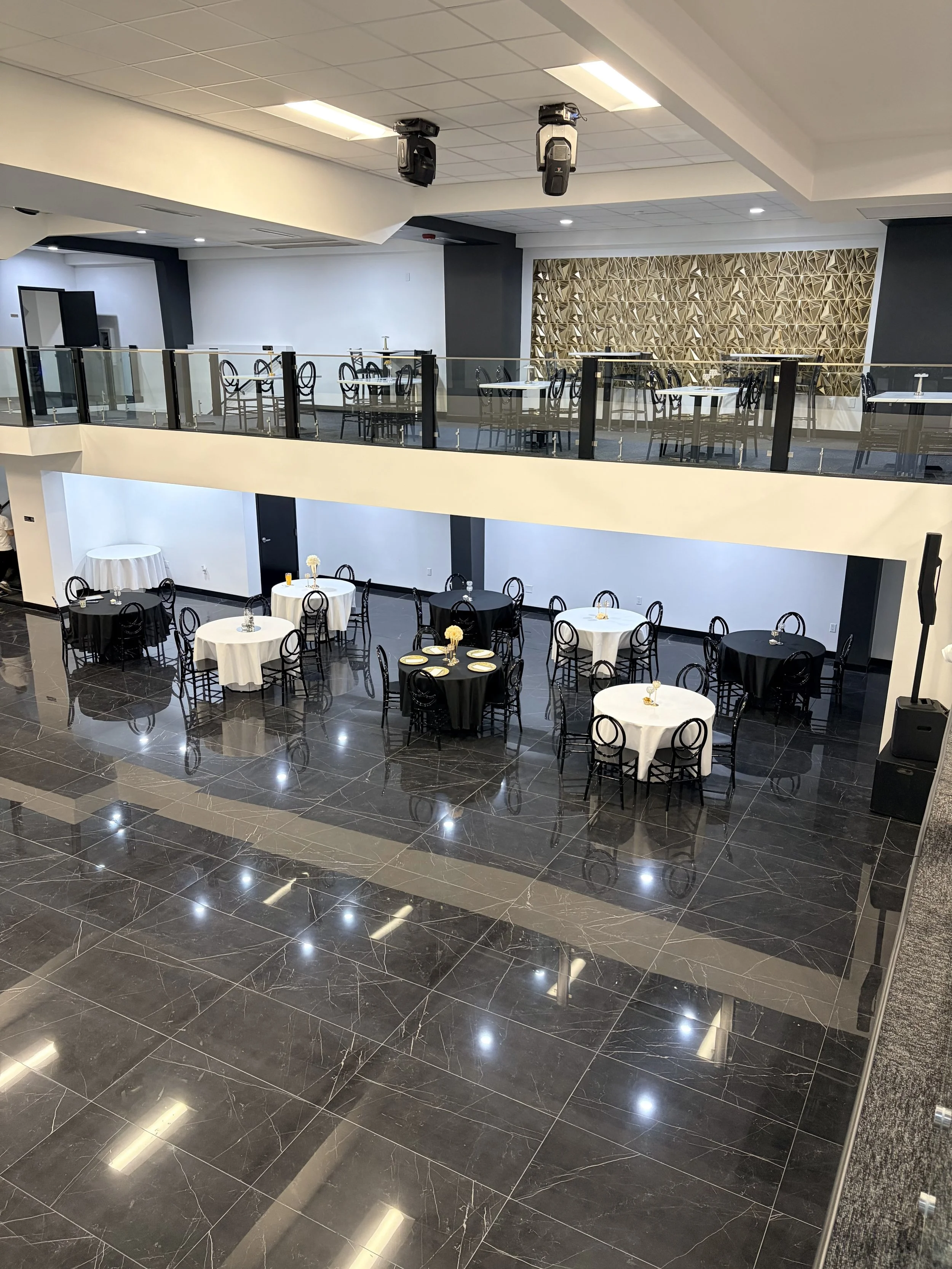An empty banquet hall or dining area with round tables, black and white tablecloths, black chairs, and a decorative wooden wall in the background. The space has glossy black marble flooring and modern lighting.