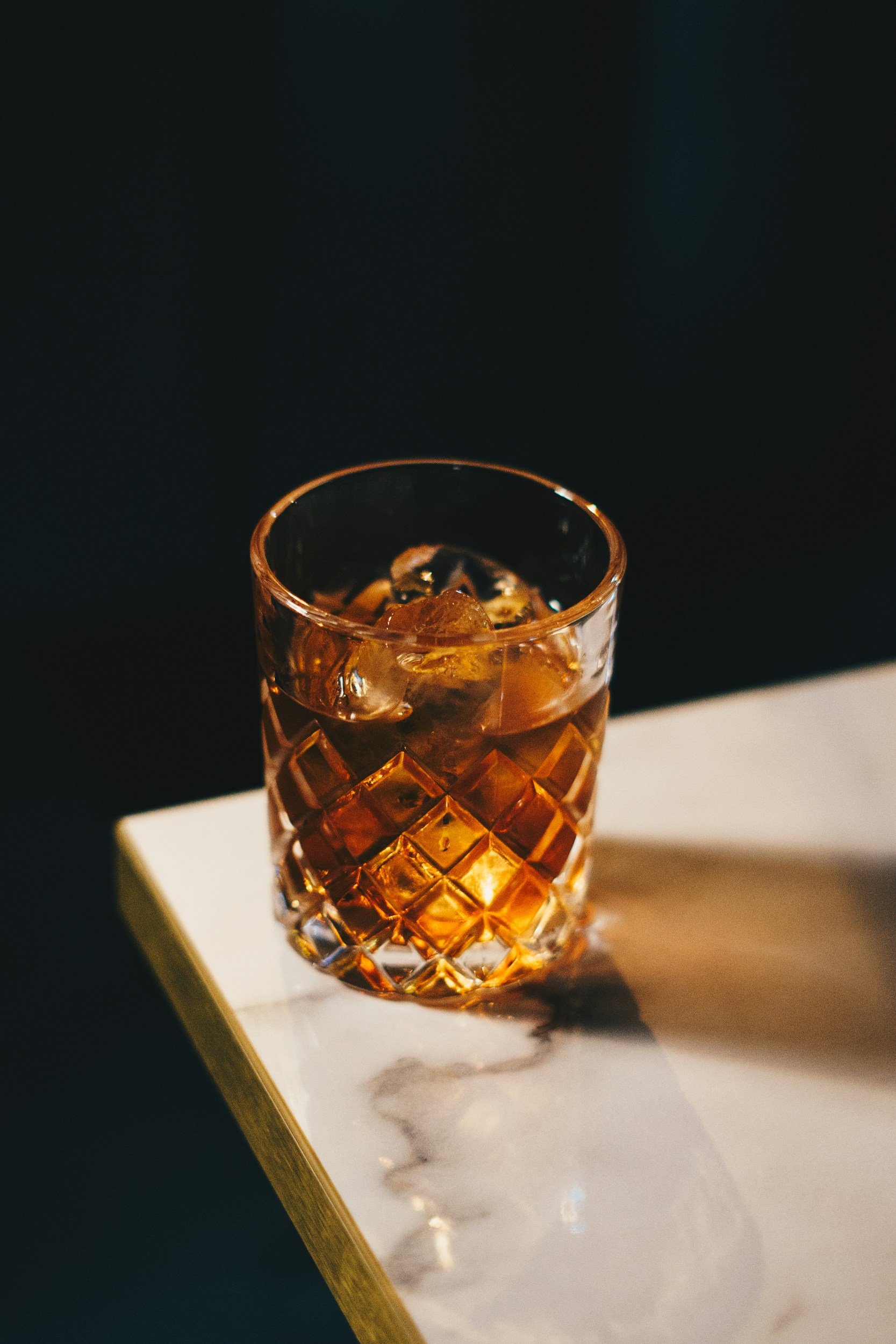 A glass of dark-colored beverage with ice cubes on a white marble surface, with a dark background.
